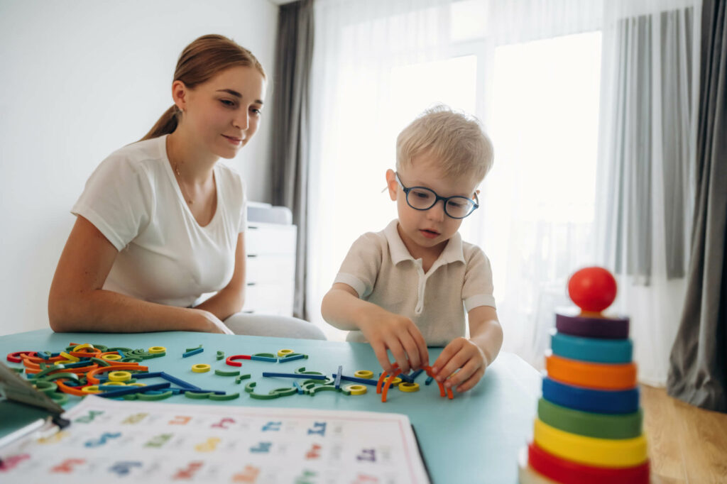 Eine Fachkraft schaut einem Kind mit Brille beim Spielen mit Lernmaterial zu